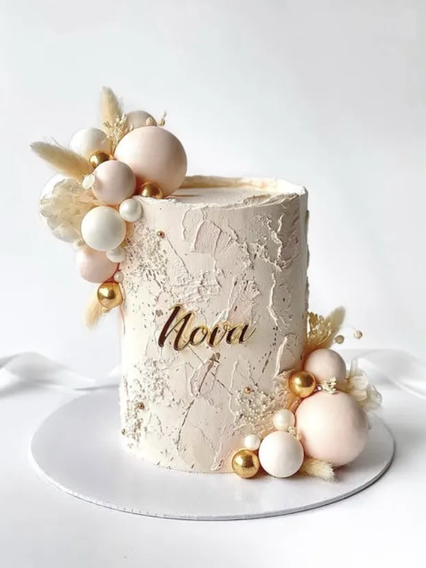 Høy hvit kake med navnet Nova i gull, pyntet med kuler i rosa, gull og hvitt